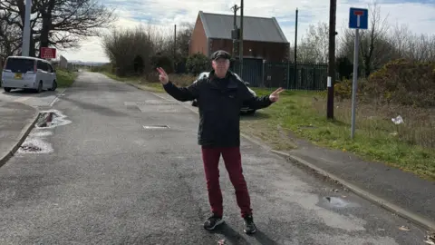Eddie Janssens A middle-aged man wearing red trousers, a black jacket and a cap stands on an empty lane smiling. He is doing peace signs with his hands. 