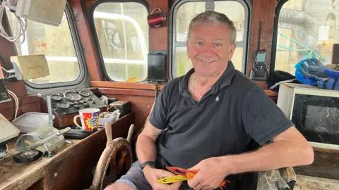 BBC/Ben Leete A man wearing a dark grey t-shirt sits inside the cabin of boat and smiles towards the camera. 