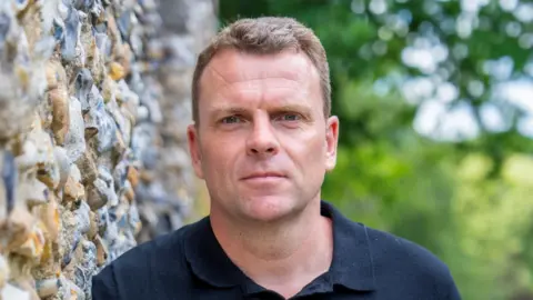 Michael Steward is wearing a navy polo top while leaning on what appears to be a harshly cobbled church wall. He has short brown hair and there is a tree behind him.