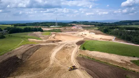 National Highways An aerial image showing the progress on the A417 Missing Link scheme. It is a large sprawling patch of land that has been levelled out, surrounded by fields and trees. In the foreground there is a digger truck with a bed full of dirt.