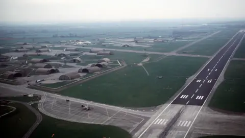 An aerial shot of a runway with green fields and low level buildings to the left.