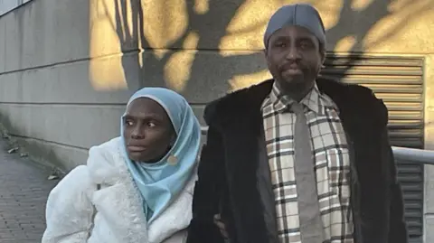 Naiyahmi and Tai Yasharahyalah outside Coventry Crown Court. Mrs Yasharahyalah is wearing a white coat and pale blue headscarf, and Mr Yasharahyalah is wearing a beige and brown shirt with a beige tie and a black coat. His head is also covered.