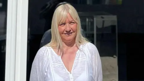 A woman with mid length blonde hair and wearing a white long sleeved top is stood i the doorway of a shop.