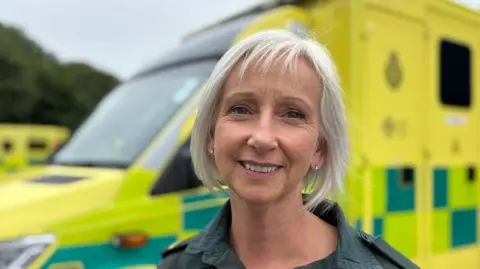 Jackie is stood outside with a yellow ambulance behind her at the ambulance station in Pembroke Dock. She is wearing the dark green ambulance service uniform. She has silvery-white hair in a bob and is smiling at the camera.