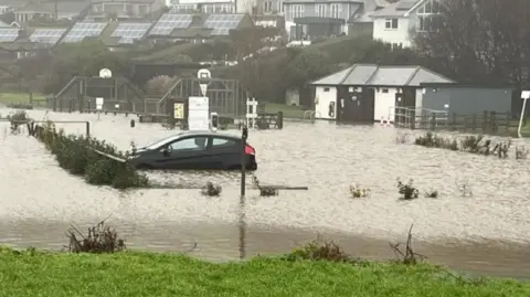 Keith Smith A black car parked in a flooded parking area. The water is covering the wheels. There is a park in the background and small buildings on the right