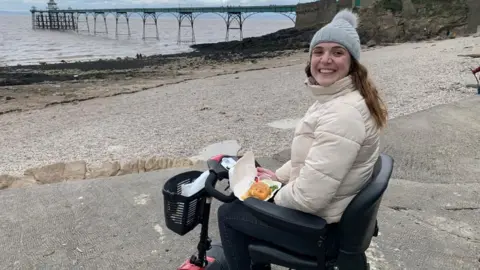 Day One Trauma Chloe Boobier is sat in a wheelchair in front of the sea, with a box of what appears to be fish and chips on her lap. She is wearing a woolly hat and a white coat.
