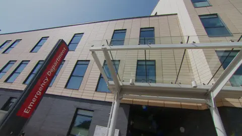 A red sign saying Emergency Department in white lettering, outside and entrance to the unit. There is a glass perspex open roof at the entrance with lots of windows visible above.