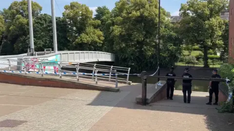 Three police officers stand by a taped-off area of river bank