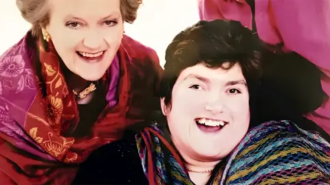 A family portrait. Alexandra has short brown hair and wears a black top and multi-coloured shawl. Rachael has short grey hair and wears a dark top and a red/pink shawl. Ian wears a smart pink shirt but his head is not visible. The women are smiling broadly.