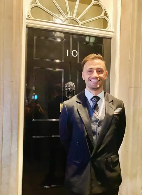 Zander  Murray Zander Murray is wearing a dark suit with a tartan tie. He is standing in front of the black door at 10 Downing Street with his hands behind his back while smiling. 