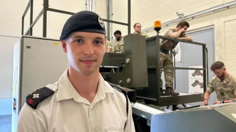 Second Lieutenant Jonathan Post stands in front of the Loader Drill Training machine, a metal simulator with platform and railings. A group of soldiers in uniform work behind him. Second Lieutenant Post wears a black beret, and white shirt with black epaulettes. 