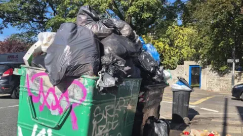 A green bin covered in graffiti overflows with black bin bags. Waste can also be seen tumbling onto the pavement of a busy city street. 