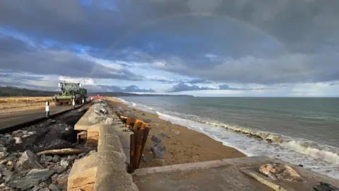 A large rainbow in the sky over a beach. The sky is dark and moody. Damage can be seen to a sea wall. A tractor is travelling along the road, which has suffered damage in a storm. Waves are lapping on the shore.