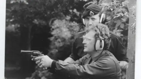 Jeff Edwards A black and white photo showing Jeff side on pointing a pistol. He wears see through goggles and large ear protectors. A man in army uniform stands next to him, with his arm on Jeff's back.