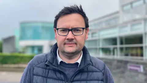 Lee Kane has short brown hair and is wearing black-rimmed glasses.
He is wearing a navy jacket, jumper and white shirt.
He is standing outside council offices in Coleraine.