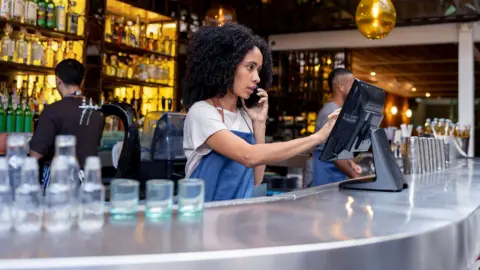 Getty Images A young woman speaks on the phone while inputting information into a point-of-sale terminal in a busy bar, with glasses on the bar next to the worker and other bar tenders in the background next to bottles on display on the shelves.