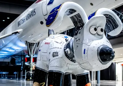 Ambitious PR A white Gromit sculpture which says Beagle 3 on it. It is seen up close in front of a British Airways plane inside a hangar.