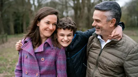 Emma Cleal A woman in a pink and purple chequered coat and a man in a dark beige puffer coat are being hugged by a boy in the middle of them who is smiling and wearing a black coat