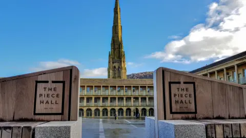 Getty Images The Piece Hall in Halifax: Large wooden benches with the words The Piece Hall on either side of the image in the middle a three-storey stone arcade is visible with a church spire behind