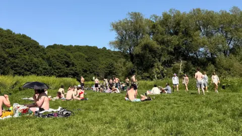 About 40 young people sitting and walking along the grass beside the River Avon in Conham River Park. The sky is clear and blue and the park is surrounded by thick woodland. Everybody is wearing bikinis, shorts or has their top off.