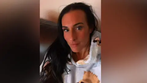 Supplied A selfie of Lisa Denton who has long dark hair and is slightly pouting into the camera. She is wearing a fluffy hoodie with dogs on them and sitting on a sofa.