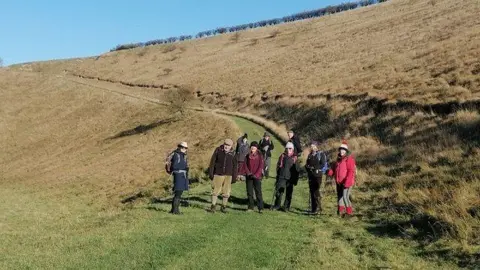 East Yorkshire Ramblers Nine ramblers facing the camera at the bottom of a grass path which curves up a gentle incline. The path has brown grass either side and a blue sky.