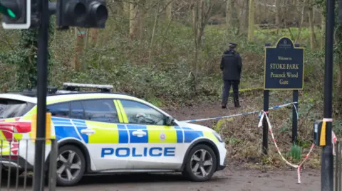 Stoke Park taped off by police tape with an officer walking into woodland and a police car by the entrance.