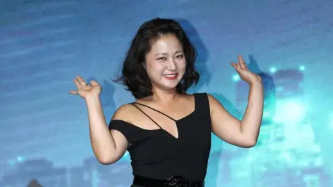 Getty Images O comediante sul-coreano Park Na-rae usa um bob preto curto e um vestido preto contra um fundo azul em um evento de imprensa.