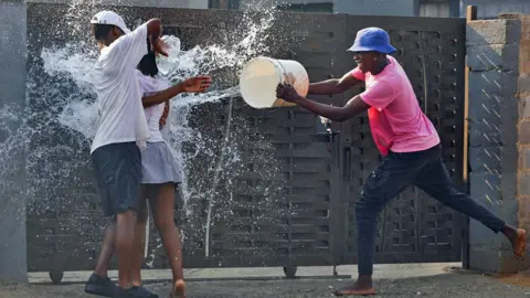 Siphiwe Sibeko/Reuters A teenager in a pink T-shirt and navy trousers with bare feet throws a bucket of water at a girl in a short skirt who has her back to the camera. Another boy pours a bottle of water over her outside a gate in Protea North, Johannesburg in South Africa - Monday 1 September 2025