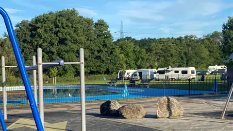 BBC A large outdoor paddling pool in a children's park. There are several camper vans and cars on the grass to the side.