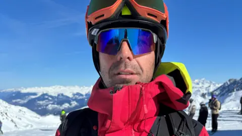 BBC/Paul Pradier A man wearing a red jacket and sunglasses and a ski helmet is seen in front of a snowy landscape