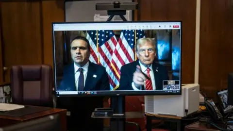 Donald Trump and his lawyer Todd Blanche (left) appear virtually on a computer desktop screen from Florida for his sentencing in his Manhattan hush money case. Trump has his finger pointing at the camera, with US flags positioned behind him and Blanche.