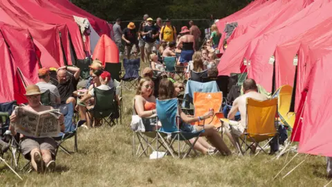 Getty Images The camera takes in a strip of grass between two rows of tents. A number of people are scattered around sitting in deckchairs and wearing summery clothes. 