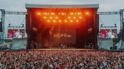 Forwards The Forwards Festival main stage lit up with orange lights at dusk in 2022. There is a large crowd of thousands of people with their arms up in the air. Little Simz is performing and her name is lit up in scrawling neon lights at the back of the stage. 