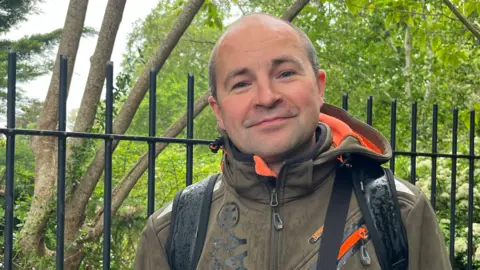 Julia Gregory/BBC A man in a dark green jacket with an orange lining, carrying a backpack. He is stood in front of a fence, through which lush greenery can be seen.