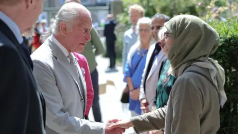King Charles III wearing a grey suit while shaking the hand of a woman at the mosque, who is wearing a smart jacket and green hijab.