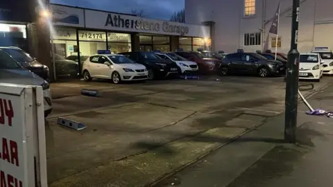 A forecourt of a garage with several cars showing and the sign "Atherstone Garage" showing. There are several empty spaces and a crumpled sign on the floor. 