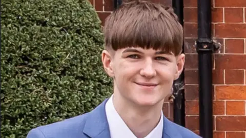 A boy with short brown hair, a blue suit jacket and white shirt, smiles as he stands in front of a brick wall, with two black drain pipes running down it, and a hedge.