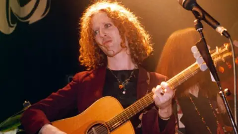 Getty Images Glen Hansard has long red, curly hair and seems to be wearing eye makeup and is playing a guitar