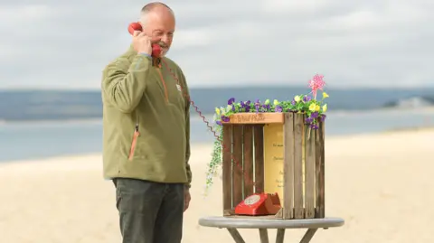 Passer-by Stephen Hoey uses a red wind phone on a table at a beach on a cloudy day. The instructions of how to use the phone are written on the box it is placed in. There are yellow and purple morning glories and a pinwheel on top of it. The sea is seen behind.