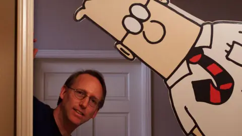 Getty Images Scott Adams and a cardboard cut-out of Dilbert, both peaking around the corner of a door frame