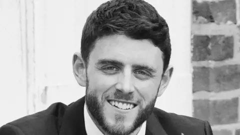 A black and white image of PC Andrew Harper a man with short brown hair and a slight beard wearing a suit and smiling at the camera.