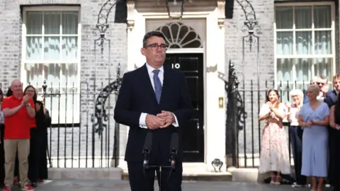 Andy Burnham wearing a suit and a blue tie, standing in front of 10 Downing Street.