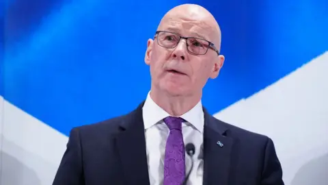 John Swinney, who is bald with glasses, speaks while standing in front of a saltire. He is wearing a business suit and purple tie 