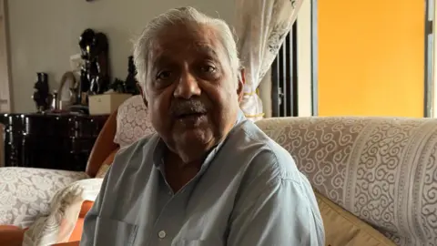 Ashraf Padanna MSR Dev looks into the camera. He is sitting on a couch in his living room. 