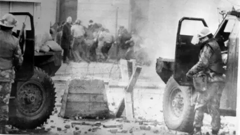PA Media File photo dated 30/01/72 of soldiers taking cover behind their sandbagged armoured cars while dispersing rioters with CS gas in Londonderry. The photo is in black and white. Barbed wire is in front of their cars and a group of people are running in the opposite direction to the soldiers. 