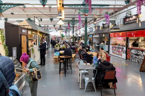 Telford & Wrekin Council An indoor market lined with stalls on either side and tables and chairs in the centre. The markets is busy with people browsing stalls and eating.