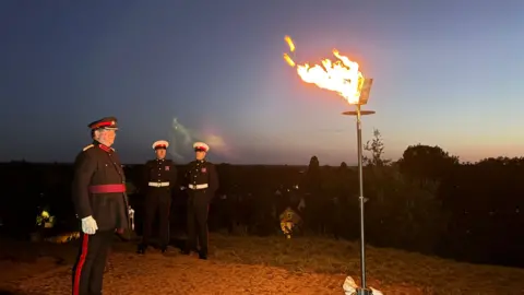 Cambridgeshire County Council Mr Christopher Walkinshaw DL (Deputy Lieutenant for Cambridgeshire) just after lighting the beacon, at the top of Castle Mound (foreground) - his military escort in background. The flame from the beacon is bright and casts a warm glow against the darkness of the evening. 