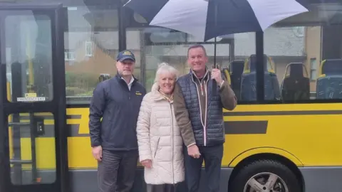 Three people stand in front of a yellow bus. One man is holding a large umbrella over the others. 