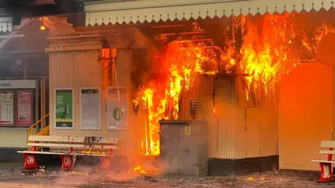 The image shows a railway station platform with part of the station building engulfed in a large fire. Bright orange flames are pouring out of the structure and through the roof, while a jet of water from firefighters can be seen spraying towards the blaze from the left-hand side, with railway tracks running across the foreground.